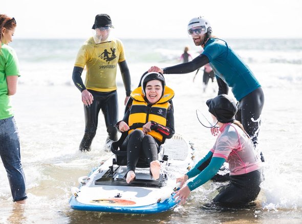 A child sits smiling in an adapted paddle board on the water with four adults around him