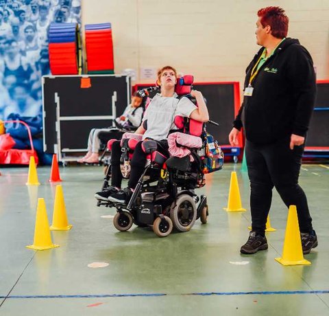 A Whizz Kidz clinician helps a young wheelchair user navigate some small cones in her powered chair