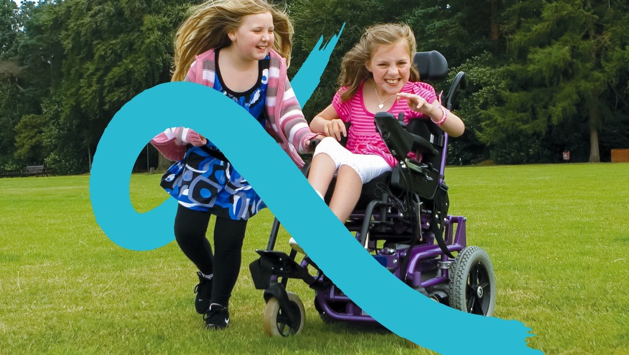 A young wheelchair user plays in her powered chair with her sister running alongside her