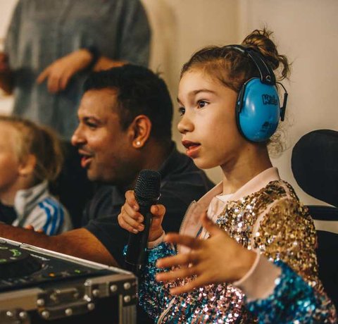 A young wheelchair user sits holding a microphone in front of a sound desk, with other people in the backgroud