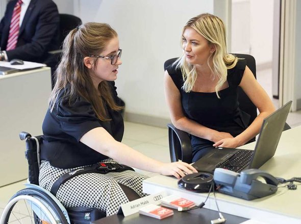 A young wheelchair user sits at a desk looking at a laptop computer with a colleague